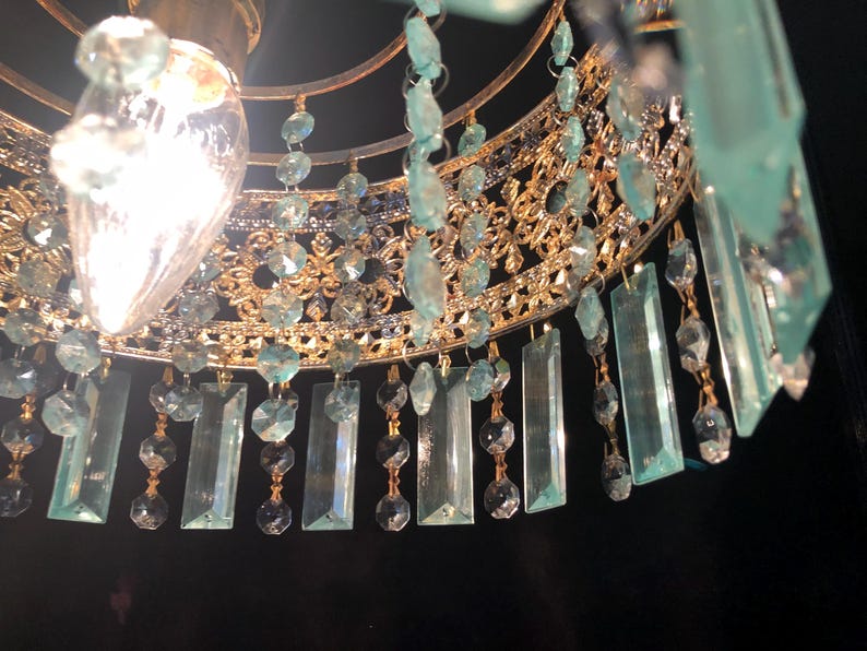 May include: Close-up of a gold-toned chandelier with intricate filigree detailing. The chandelier features hanging rectangular and round aquamarine-colored crystals. A bright light bulb is visible at the top, illuminating the crystals and gold frame.