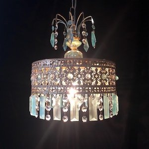 May include: An ornate chandelier with a bronze-colored metal frame and a cylindrical shade. The shade features intricate floral cutouts and is adorned with clear and light blue crystal accents. The chandelier hangs from a chain and emits a warm glow.