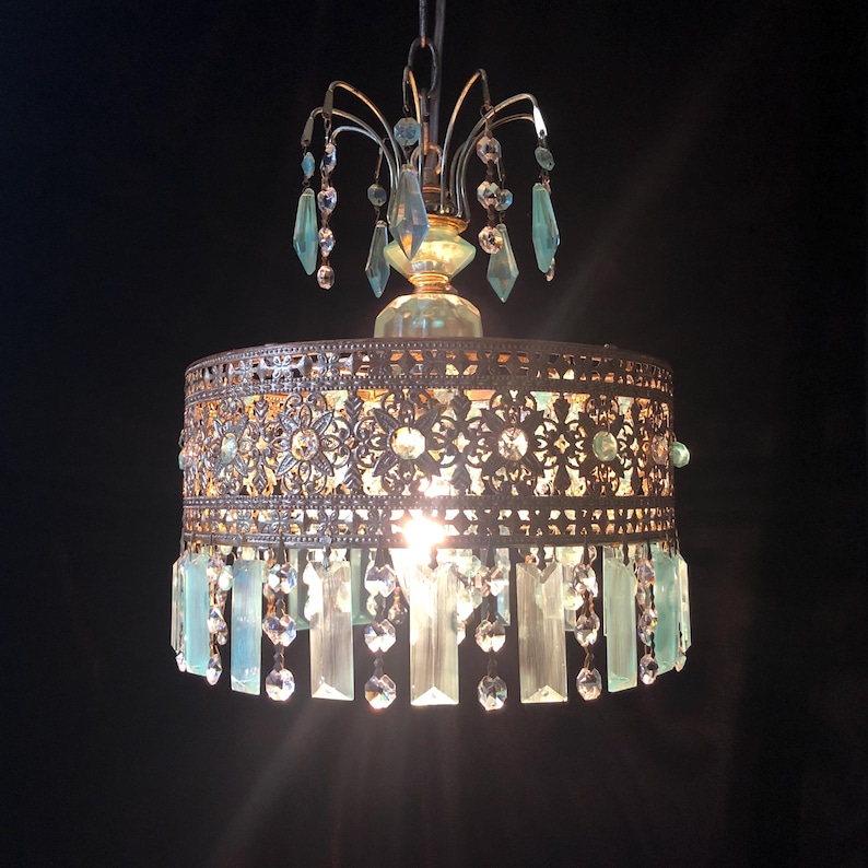 May include: An ornate chandelier with a bronze-colored metal frame and a cylindrical shade. The shade features intricate floral cutouts and is adorned with clear and light blue crystal accents. The chandelier hangs from a chain and emits a warm glow.
