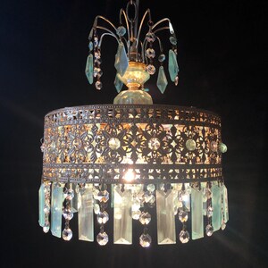 May include: An antique-style chandelier with a bronze-colored metal frame and intricate cut-out designs. The chandelier features hanging crystal accents and light blue glass pendants, creating a vintage aesthetic. The light fixture hangs from a chain.