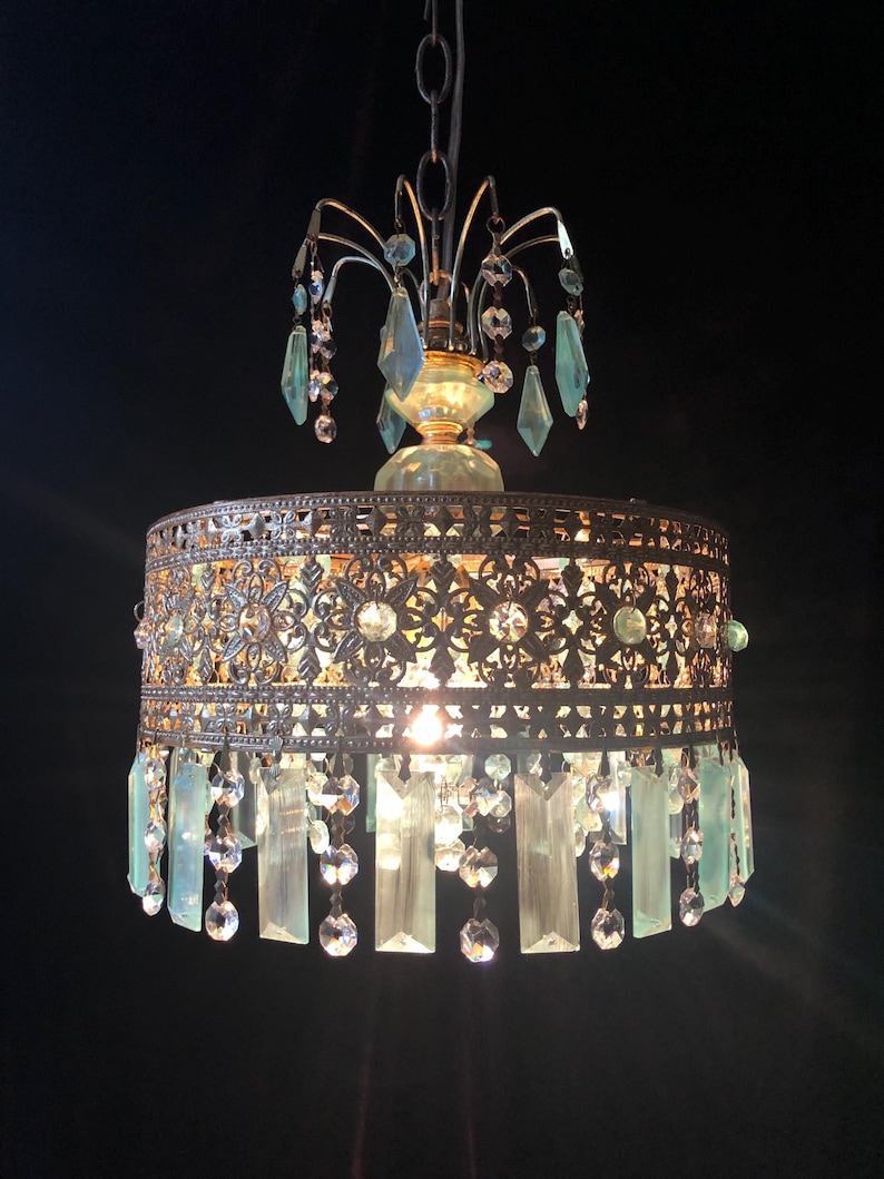 May include: An antique-style chandelier with a bronze-colored metal frame and intricate cut-out designs. The chandelier features hanging crystal accents and light blue glass pendants, creating a vintage aesthetic. The light fixture hangs from a chain.