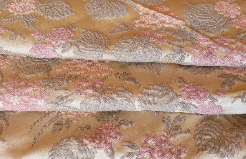 May include: Close-up of a fabric with a floral pattern. The fabric is a light beige color with a raised texture. The flowers are pink and gray and have a delicate, feminine look.