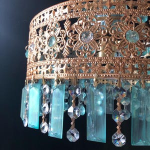 May include: Close-up of a vintage chandelier with a gold-colored filigree frame. The frame features floral and geometric cutouts, and is adorned with turquoise and clear crystal accents. Hanging below are rectangular turquoise crystals and clear crystal beads.