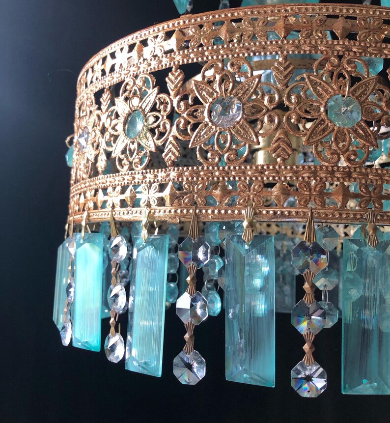 May include: Close-up of a vintage chandelier with a gold-colored filigree frame. The frame features floral and geometric cutouts, and is adorned with turquoise and clear crystal accents. Hanging below are rectangular turquoise crystals and clear crystal beads.
