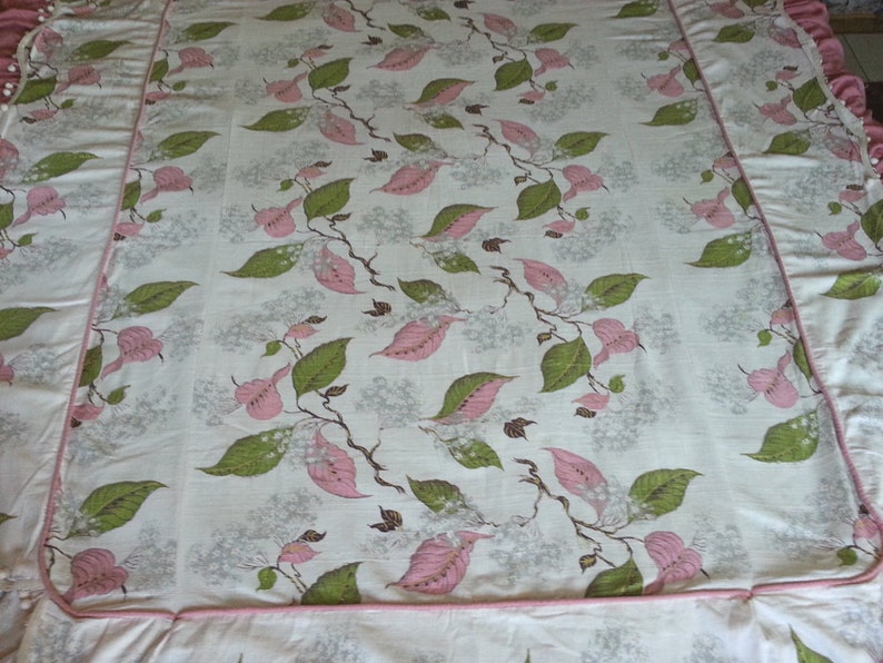 May include: A white bedspread with a pink and green floral pattern. The bedspread has a pink ruffled border.