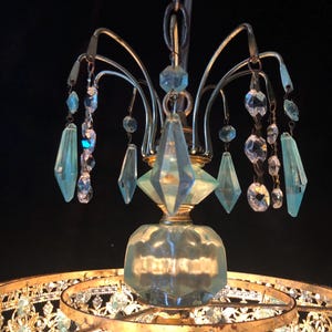 May include: A vintage chandelier with a gold-toned metal frame and hanging crystal accents. The chandelier features clear and light blue crystal pendants and a decorative glass base. The design is ornate and elegant, suitable for interior decor.