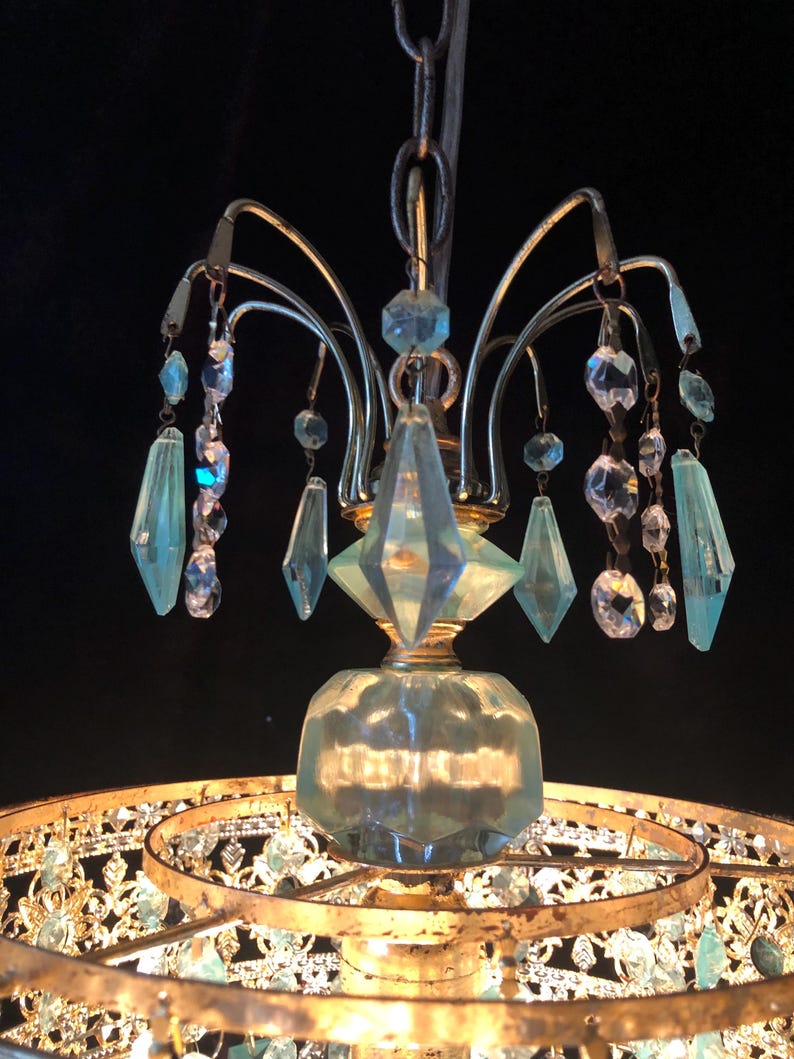 May include: A vintage chandelier with a gold-toned metal frame and hanging crystal accents. The chandelier features clear and light blue crystal pendants and a decorative glass base. The design is ornate and elegant, suitable for interior decor.