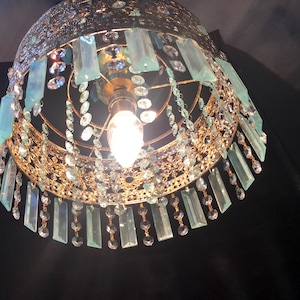 May include: Ornate gold-toned chandelier with a domed shape. The fixture is adorned with hanging rectangular blue-green crystals and smaller clear crystals. A single light bulb is visible at the center, illuminating the intricate design. The chandelier is suspended against a dark background.