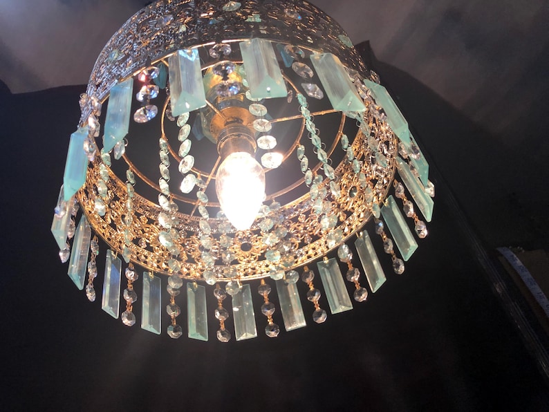 May include: Ornate gold-toned chandelier with a domed shape. The fixture is adorned with hanging rectangular blue-green crystals and smaller clear crystals. A single light bulb is visible at the center, illuminating the intricate design. The chandelier is suspended against a dark background.