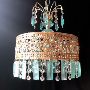 May include: A vintage-style chandelier with a gold-colored filigree shade and hanging blue-green and clear crystal accents. The chandelier has a decorative floral pattern and is suspended from a chain.