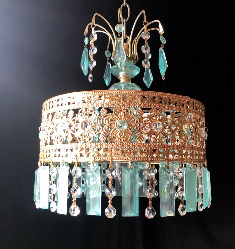 May include: A vintage-style chandelier with a gold-colored filigree shade and hanging blue-green and clear crystal accents. The chandelier has a decorative floral pattern and is suspended from a chain.