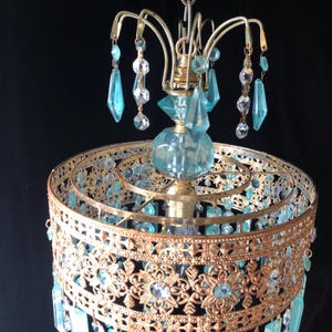 May include: Vintage chandelier with a gold-toned metal frame and ornate filigree detailing. It features hanging blue and clear crystal accents and a central blue glass element. The chandelier is suspended against a dark background.