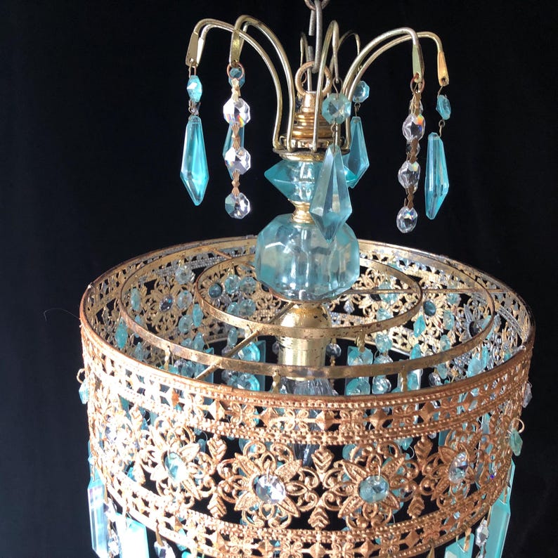 May include: Vintage chandelier with a gold-toned metal frame and ornate filigree detailing. It features hanging blue and clear crystal accents and a central blue glass element. The chandelier is suspended against a dark background.