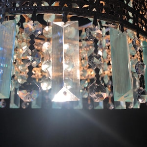May include: Close-up of a decorative chandelier with a dark metal frame. The chandelier features clear and light blue crystal accents, including rectangular and faceted shapes, hanging from the frame. The crystals reflect light, creating a sparkling effect.