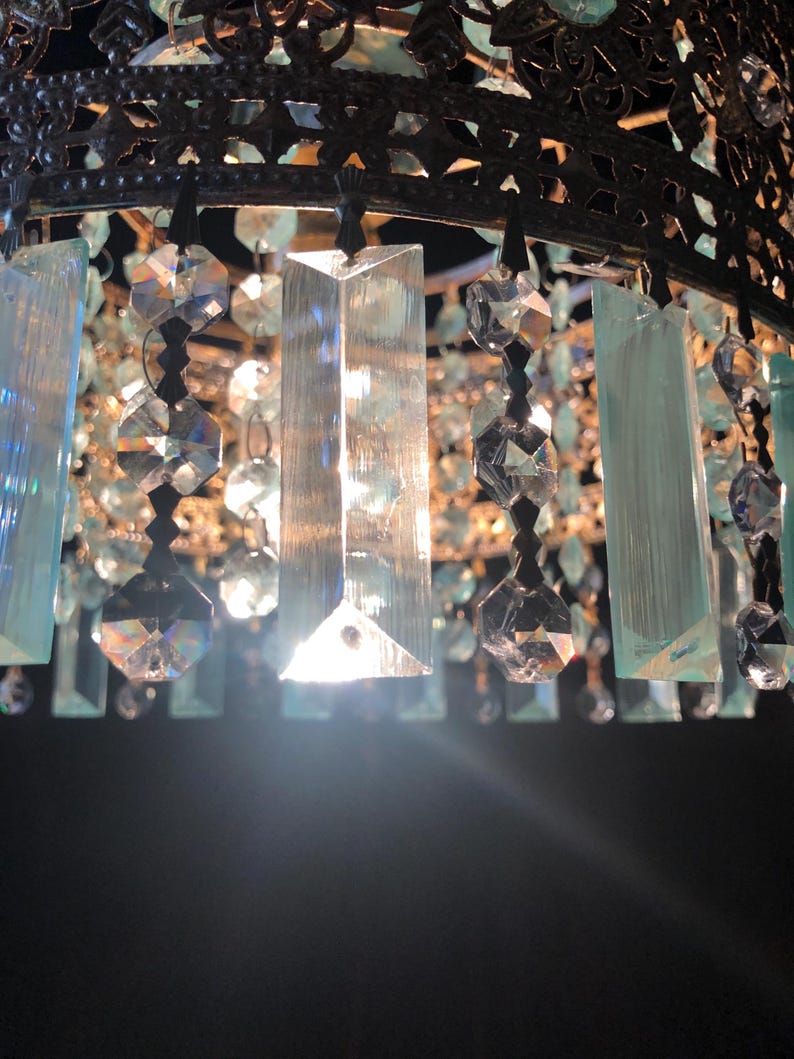 May include: Close-up of a decorative chandelier with a dark metal frame. The chandelier features clear and light blue crystal accents, including rectangular and faceted shapes, hanging from the frame. The crystals reflect light, creating a sparkling effect.
