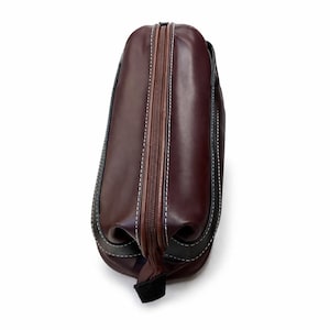 May include: A brown leather toiletry bag with a dark brown zipper and white stitching. The bag has black trim and a black fabric loop. The bag is rectangular with rounded edges and is set against a white background.