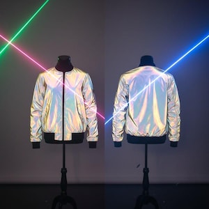 Puede incluir: Chaqueta bomber iridiscente exhibida en un maniquí, vistas frontal y posterior. La chaqueta tiene un acabado reflectante que cambia de color con ribetes negros en el cuello, los puños y el dobladillo. La chaqueta está cerrada con cremallera. El fondo presenta rayos láser verdes, rosas y azules.