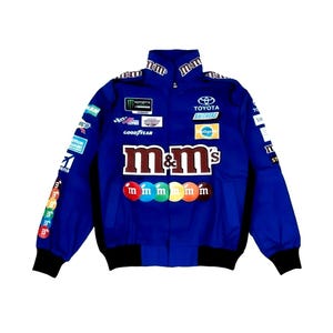 May include: A blue racing jacket with black trim, featuring sponsor logos and the M&M's logo on the front. The jacket has a high collar and a zip-up front. The sleeves have colorful graphics. The jacket is a vibrant piece of apparel.