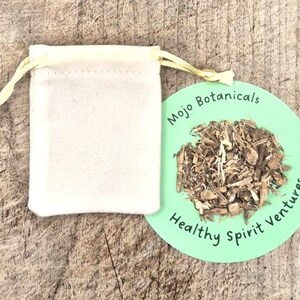 Puede incluir: Una pequeña bolsa con cordón de color blanquecino junto a una etiqueta circular verde con el texto "Mojo Botanicals Healthy Spirit Ventures". La etiqueta muestra un montón de material vegetal marrón seco. Los artículos descansan sobre una superficie de madera envejecida.