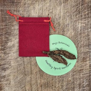 Puede incluir: Una bolsa roja con cordón y una etiqueta circular verde claro con el texto "Mojo Botanicals" y "Healthy Spirit Ventures". Dos elementos botánicos marrones secos están en la etiqueta. Los artículos están sobre una superficie de madera.