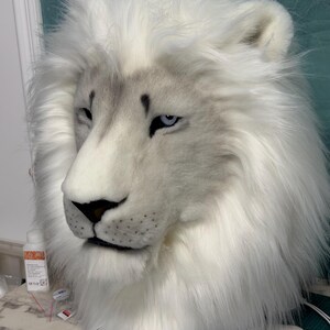 May include: A white lion mask with a detailed, realistic design. The mask features a fluffy white mane, a gray muzzle, and piercing blue eyes. The lion's nose is black, and the fur appears soft and plush.