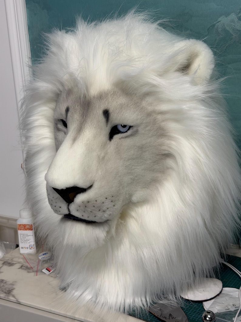 May include: A white lion mask with a detailed, realistic design. The mask features a fluffy white mane, a gray muzzle, and piercing blue eyes. The lion's nose is black, and the fur appears soft and plush.