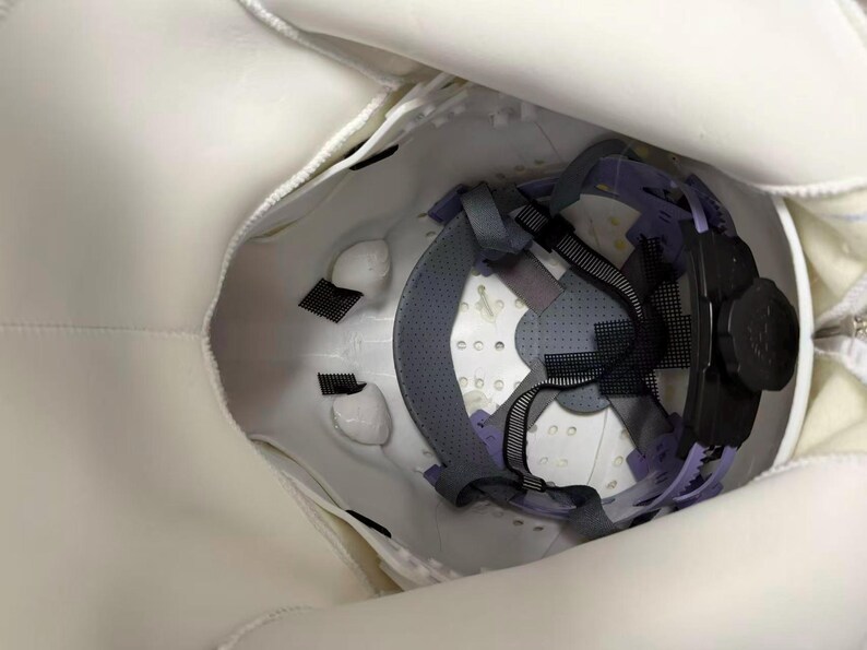May include: White safety helmet with interior suspension system. The interior features a gray and purple harness, black straps, and a black adjustment knob. The helmet is designed for head protection.