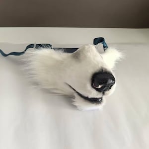 Könnte beinhalten: Eine weiße und schwarze, pelzige Wolfsmaske mit schwarzer Nase und Mund. Die Maske hat einen blauen Riemen. Die Maske besteht aus weichem, plüschigem Material.