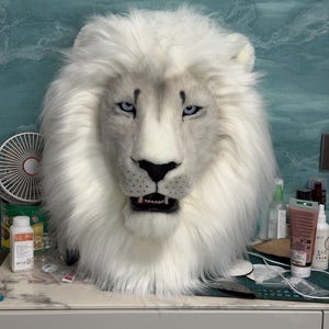 May include: A white lion mask with a fluffy mane and detailed facial features. The mask has blue eyes, a black nose, and an open mouth revealing teeth. The mask is likely for costume or cosplay purposes.