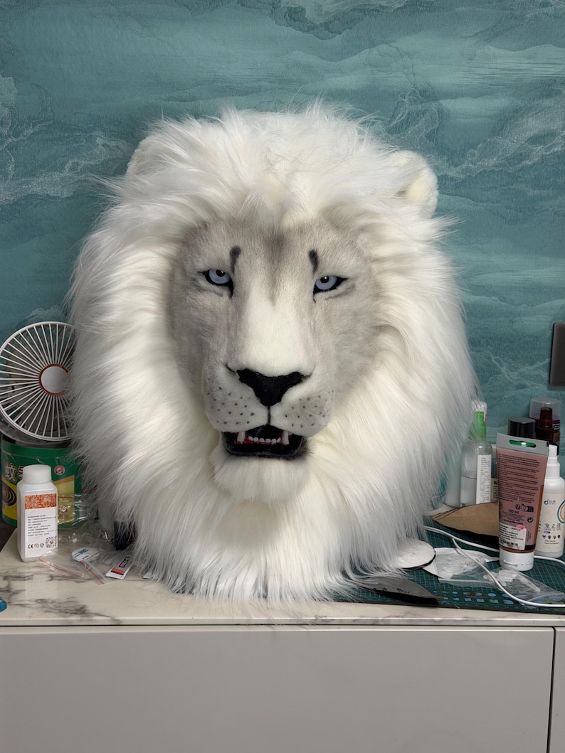 May include: A white lion mask with a fluffy mane and detailed facial features. The mask has blue eyes, a black nose, and an open mouth revealing teeth. The mask is likely for costume or cosplay purposes.