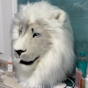 May include: A white lion mask with a realistic design. The mask features a fluffy mane, detailed facial features, and blue eyes. The mask is made of soft, plush material and is likely for costume or cosplay use.