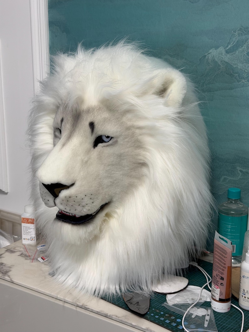 May include: A white lion mask with a realistic design. The mask features a fluffy mane, detailed facial features, and blue eyes. The mask is made of soft, plush material and is likely for costume or cosplay use.
