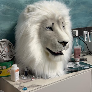 May include: A white lion mask with blue eyes and a detailed, realistic design. The mask features a full mane of fluffy white fur and is likely intended for costume or cosplay use. The lion's expression is neutral, with a slightly open mouth.