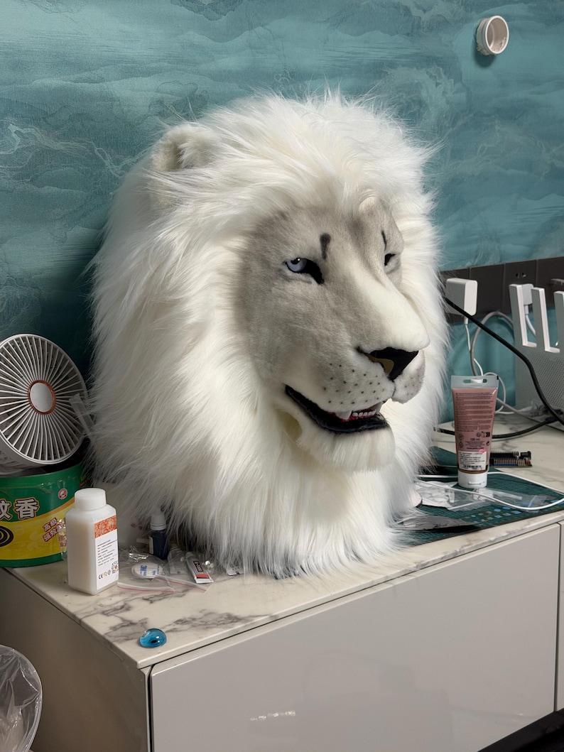 May include: A white lion mask with blue eyes and a detailed, realistic design. The mask features a full mane of fluffy white fur and is likely intended for costume or cosplay use. The lion's expression is neutral, with a slightly open mouth.