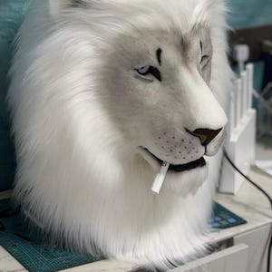 May include: A white lion mask with a detailed design. The mask features a realistic face with blue eyes, a brown nose, and a white mane. A white object is in the lion's mouth. The mask is likely for cosplay or costume use.