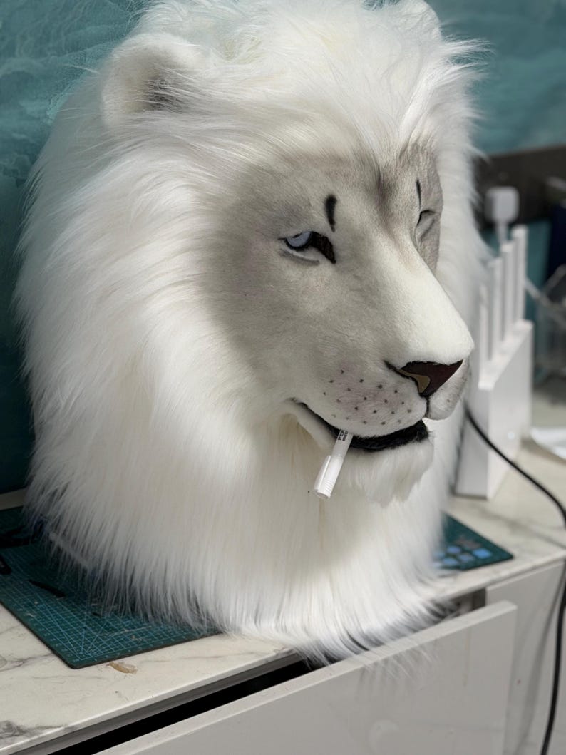 May include: A white lion mask with a detailed design. The mask features a realistic face with blue eyes, a brown nose, and a white mane. A white object is in the lion's mouth. The mask is likely for cosplay or costume use.