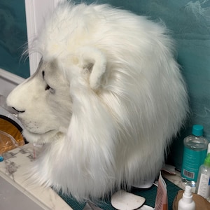 May include: A white lion mask with a fluffy mane and detailed facial features. The mask is made of soft, plush material and has a realistic appearance. The lion's nose and ears are visible, and the mask is designed to be worn on the head.