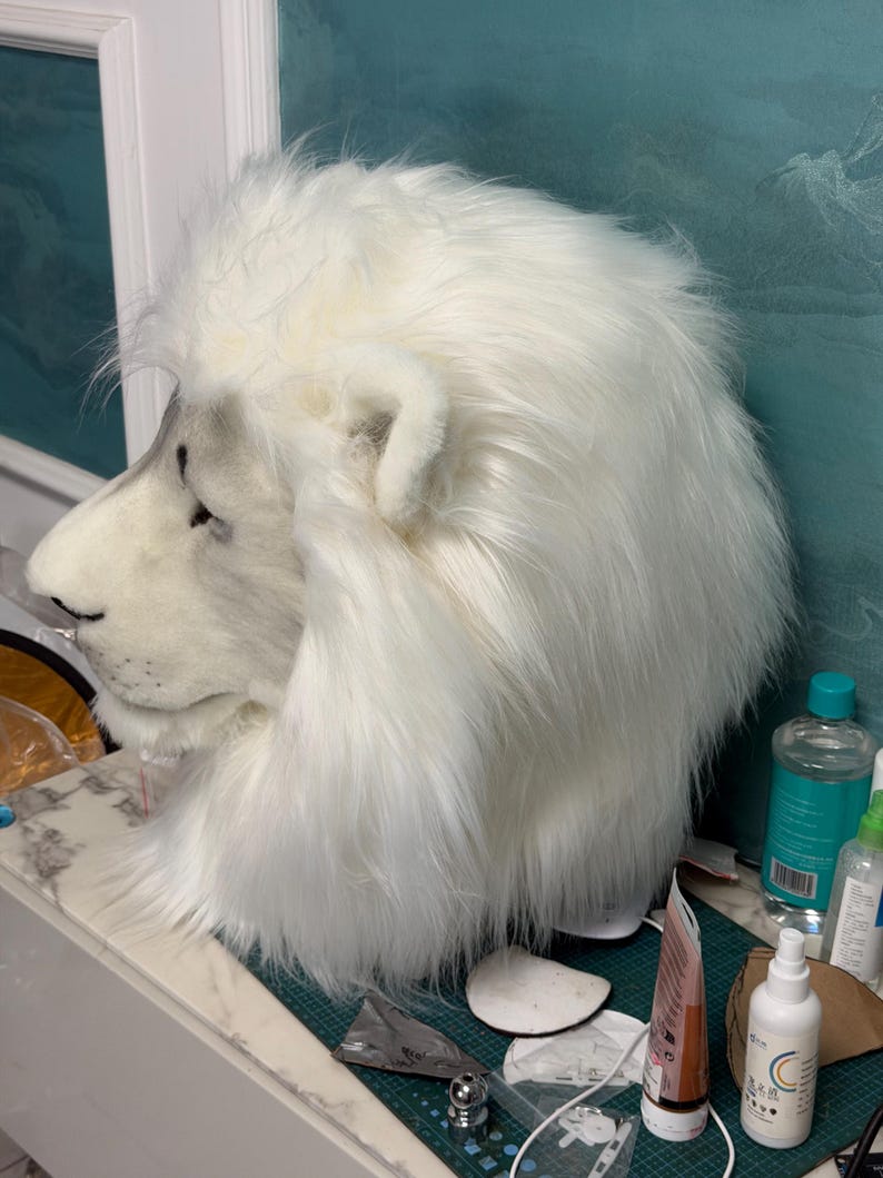 May include: A white lion mask with a fluffy mane and detailed facial features. The mask is made of soft, plush material and has a realistic appearance. The lion's nose and ears are visible, and the mask is designed to be worn on the head.