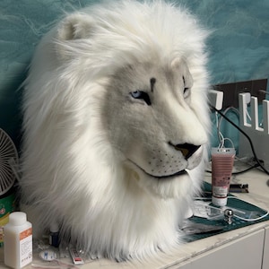 May include: A white lion mask with a realistic design. The mask features a fluffy mane, a detailed face with blue eyes, and a black nose. The mask is made of soft, plush material, and is likely for costume or cosplay use.