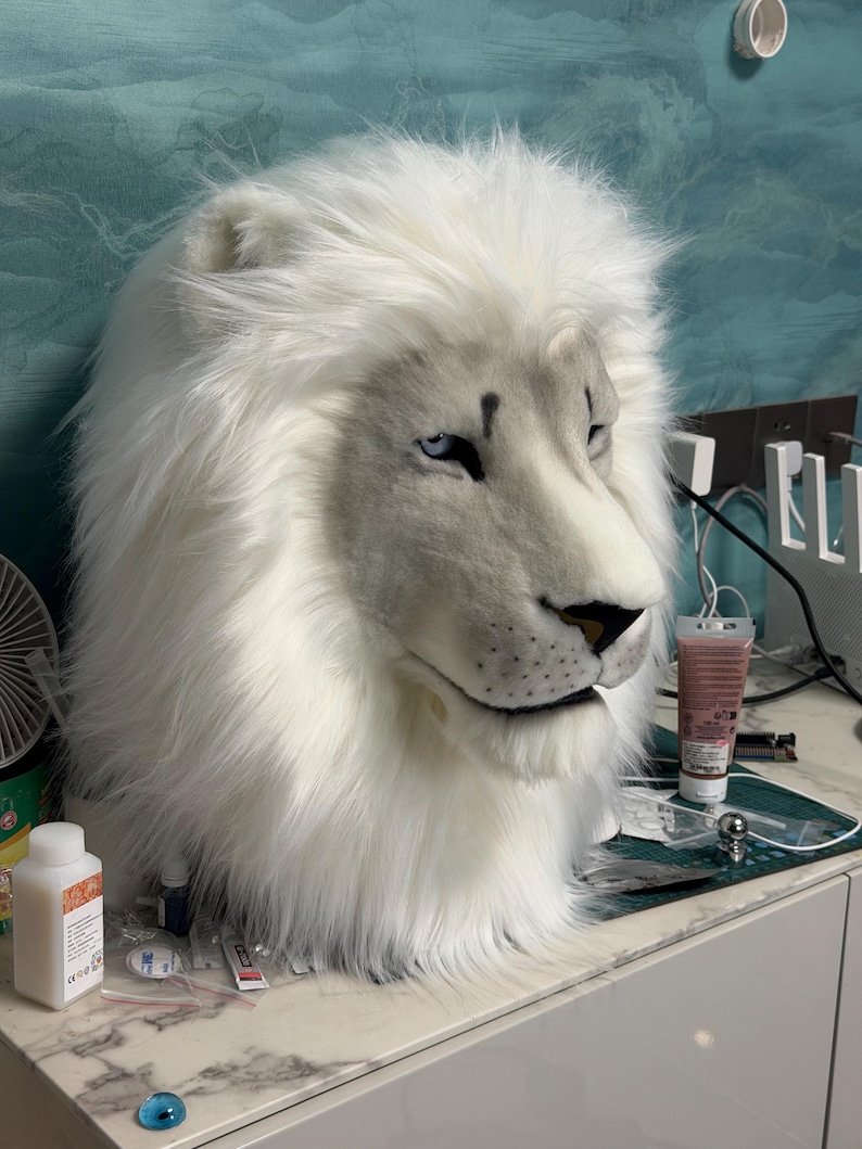 May include: A white lion mask with a realistic design. The mask features a fluffy mane, a detailed face with blue eyes, and a black nose. The mask is made of soft, plush material, and is likely for costume or cosplay use.