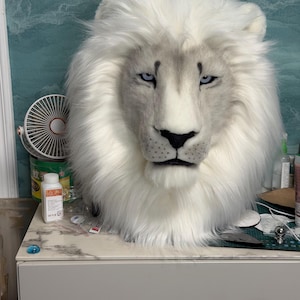 May include: A white lion mask with a fluffy mane and piercing blue eyes. The mask has a realistic design with detailed facial features, including a black nose and subtle gray shading.