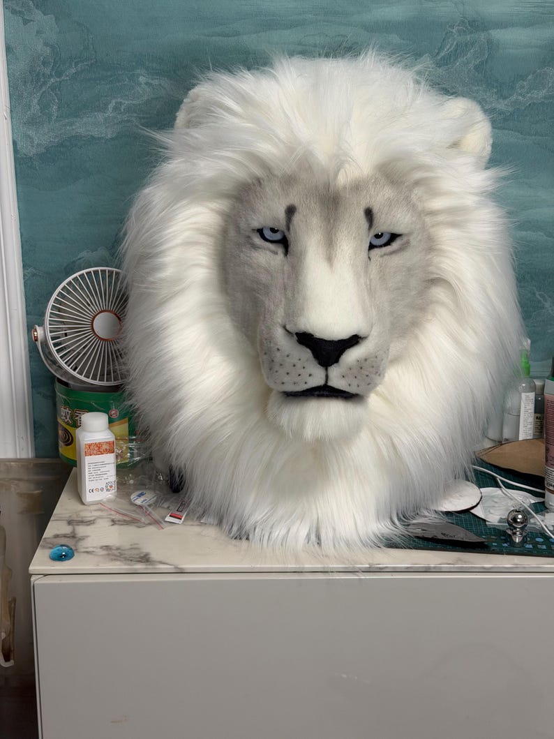May include: A white lion mask with a fluffy mane and piercing blue eyes. The mask has a realistic design with detailed facial features, including a black nose and subtle gray shading.