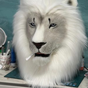 May include: A white lion mask with a stern expression, featuring a full mane and detailed facial features. The mask has a cigarette in its mouth. The background is a light teal color.