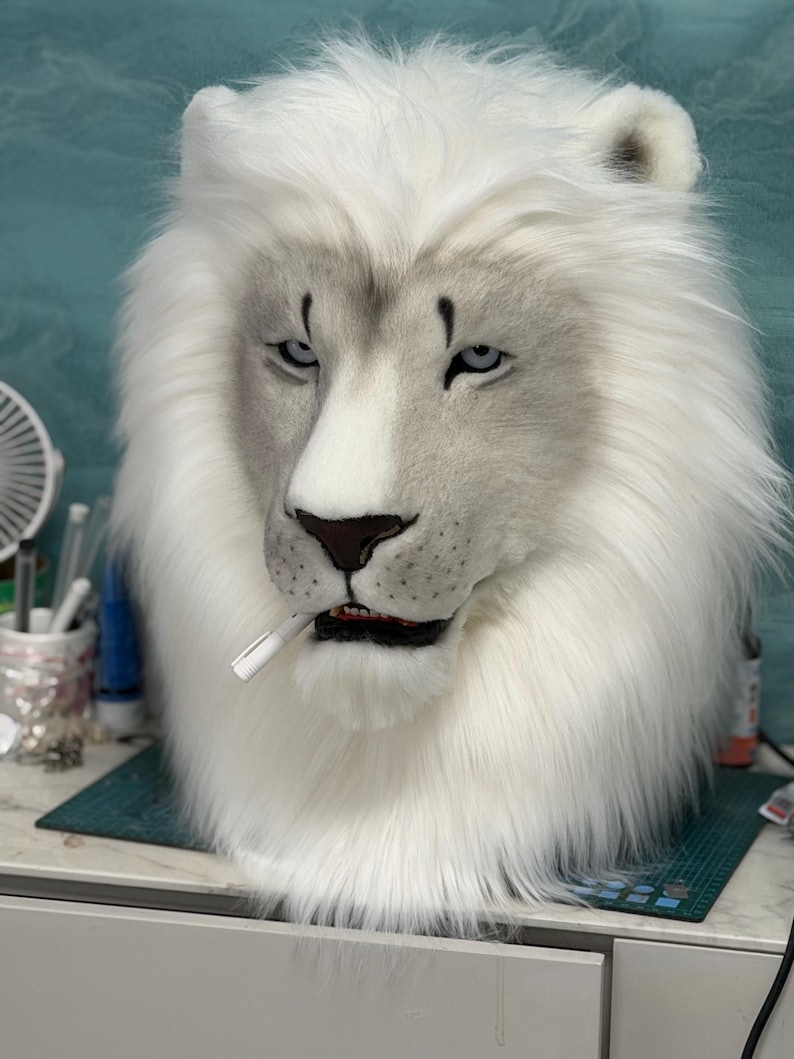 May include: A white lion mask with a stern expression, featuring a full mane and detailed facial features. The mask has a cigarette in its mouth. The background is a light teal color.