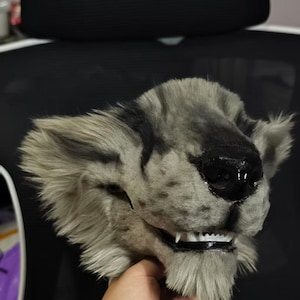Könnte beinhalten: Eine detaillierte, pelzige Maske einer katzenartigen Kreatur mit einem grau-schwarzen Farbschema. Die Maske hat ein realistisches Design mit sichtbaren Zähnen und einer schwarzen Nase. Die Ohren sind aufrecht und das Fell wirkt weich und strukturiert.