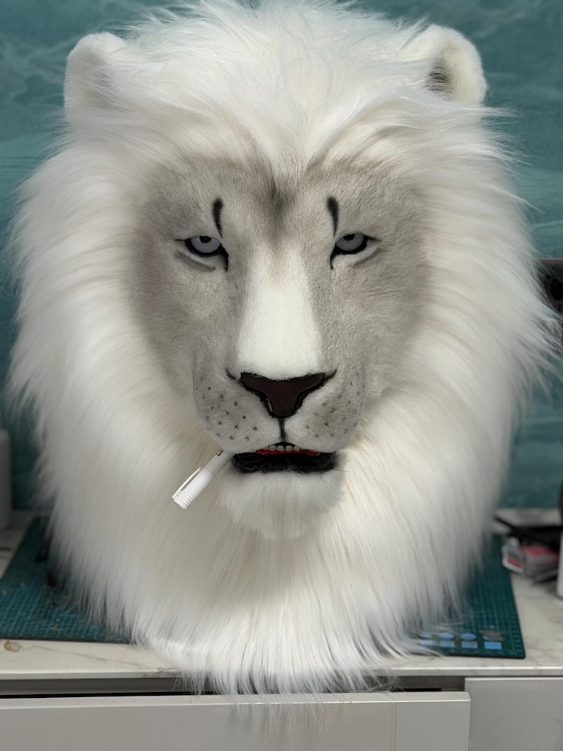 May include: A white lion mask with a realistic design. The mask features a full mane of fluffy white fur, gray accents around the eyes and nose, and a small white object in its mouth. The lion's expression is serious.