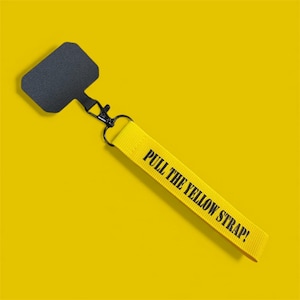 Puede incluir: Una correa de teléfono amarilla con el texto "PULL THE YELLOW STRAP!" en negro. La correa está conectada a un soporte de teléfono negro y a un cierre de metal negro. El fondo es amarillo liso.