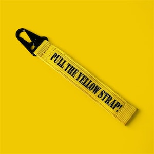 May include: Yellow strap with the text "PULL THE YELLOW STRAP!" in black. It features a black metal clip. The background is a solid yellow colour.