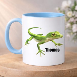 Könnte beinhalten: Weiße Keramiktasse mit hellblauem Innenraum und Henkel. Die Tasse zeigt eine detaillierte Illustration einer grünen Eidechse mit dem Namen "Thomas" in Schwarz. Die Tasse steht auf einer Holzoberfläche.