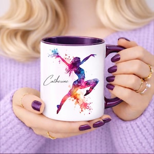 Peut inclure: Mug en céramique blanche avec intérieur et anse violets. Le mug présente un motif aquarelle coloré d'une danseuse avec le nom "Catherine" en écriture cursive. La danseuse est représentée dans des tons de rose, bleu et orange.